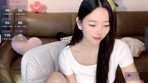 Media: Video of an East Asian woman with long black hair, wearing a white T-shirt, sitting on a beige couch with stuffed animals in a cozy, indoor setting.