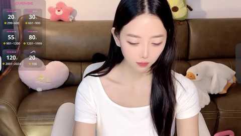 Media: Video of an Asian woman with long black hair, wearing a white shirt, sitting on a brown leather couch. Background includes plush toys, a digital clock, and a virtual reality display.