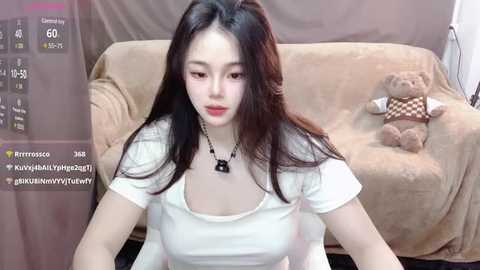 Media: Video of a young Asian woman with long black hair, wearing a white top, seated on a beige couch. Background features a teddy bear and a gray curtain.