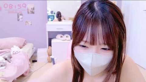 Media: Video of an Asian woman with straight brown hair and a surgical mask, sitting in a pastel-colored bedroom. The background shows a bed, white furniture, and a wall adorned with small posters.