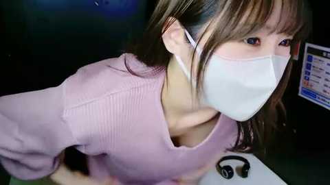 Media: Video of an Asian woman with straight brown hair, wearing a white surgical mask, pink ribbed sweater, and white headphones, leaning forward on a desk with a laptop and black headphones.