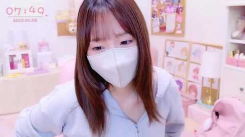 Media: A video of an East Asian woman with long brown hair, wearing a white face mask, light blue top, and pink jacket, sitting in a pastel-colored, organized room with shelves and posters.