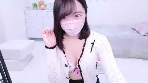 Media: A video of an East Asian woman with shoulder-length black hair and a light complexion, wearing a white lace cardigan, pink bra, and pink face mask, in a minimalist, white bedroom with a bed and dresser.