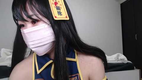 Media: Video of a woman in a traditional Chinese dress with a white surgical mask, long black hair, and a red hair ornament. She stands in a simple, minimally decorated room with a white bed and a dark wardrobe in the background.