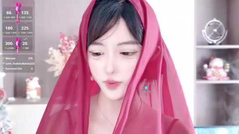 Media: Video of a young Asian woman in a red veil, standing indoors. Background features a shelf with decorative items and a blurred view of a room.