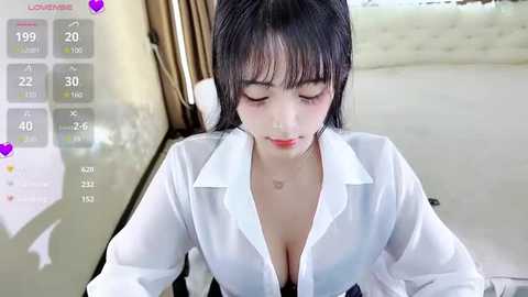 Media: A video of an Asian woman with straight black hair, wearing a white blouse with a deep neckline, in a modern bedroom setting.