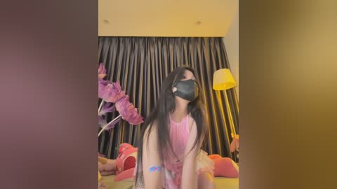 Media: Video of a young woman with long black hair, wearing a pink dress and face mask, sitting on a bed with pink and red bedding, against dark drapes and a yellow lamp.