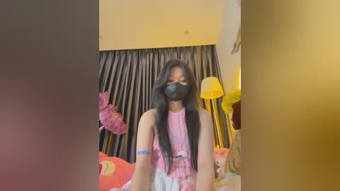 Media: Video of an Asian woman with long black hair and light skin, wearing a pink sleeveless top, black face mask, and blue wristband, standing in a bedroom with a yellow lamp, pink teddy bear, and dark curtains.