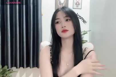 Media: Video of a young Asian woman with long black hair, fair skin, and red lipstick, wearing a sheer top, sitting on a couch. Background features a framed photo and dark curtains.