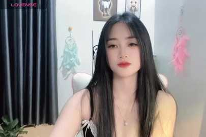 Media: A video of an East Asian woman with long black hair, wearing a yellow dress, standing in a softly lit room with framed pictures on the wall and hanging clothes.