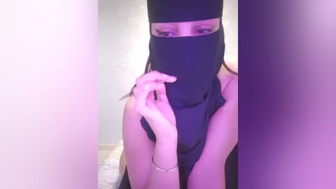 Media: A video of a woman with light skin wearing a black burqa, partially covering her face and head, in a dimly lit room with purple lighting.