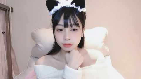 Media: A video of an East Asian woman with long black hair, wearing a white off-shoulder sweater, black cat ears headband, and white frilly collar, seated on a beige cushion, smiling softly.