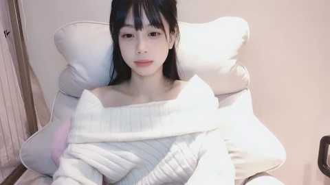 Media: Video of an Asian woman with straight black hair and pale skin, wearing a white off-shoulder sweater, lounging on a white leather chair with a quilted blanket. The background is a plain beige wall.