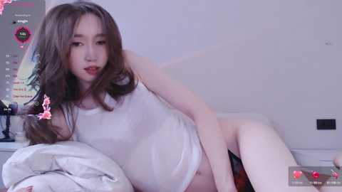 A video of an Asian woman with long brown hair, fair skin, and red lipstick, wearing a white tank top and black shorts, reclining on a bed, with a pink toy on the pillow.