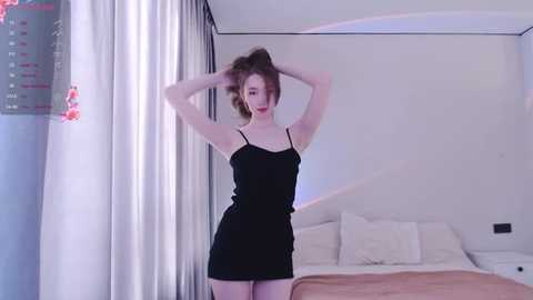 Media: Video of a fair-skinned, slim young woman with brown hair in a messy bun, wearing a black spaghetti-strap dress, posing in a modern bedroom with white walls and a neatly made bed.