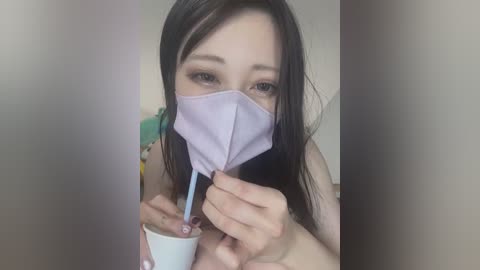 Media: Video of a young Asian woman with long, straight black hair, wearing a light purple face mask, holding a white disposable cup with a blue straw. She's indoors with blurred background.