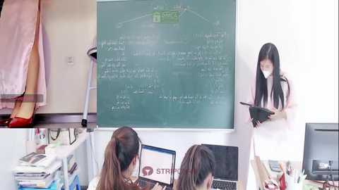 Media: Video of a classroom with two students facing a large green chalkboard, one wearing a pink dress, the other in a white dress. The teacher, in a white dress, stands at the board.