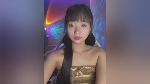 Media: Video of a young Asian woman with long black hair, bangs, and light skin, wearing a strapless, beige dress with a floral pattern, standing in a dimly lit room with colorful, abstract wall art in the background.