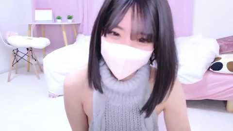 Media: Video of an Asian woman with straight black hair, wearing a white face mask, grey turtleneck sweater, and a pink turtleneck sweater, in a pastel-themed bedroom with pink walls and furniture.