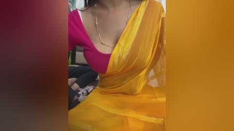 Media: A close-up video of a woman wearing a bright yellow saree over a pink blouse, with a delicate gold necklace. Her face is not visible. The background is blurred, featuring a green object.