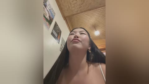 Media: A video of an Asian woman with long dark hair, wearing a white spaghetti-strap top, looking up with a serene expression, standing in a cozy room with wooden ceiling and framed pictures on the wall.