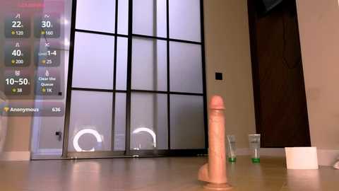 Video of a minimalist modern room with a tall, realistic-looking pink dildo on a beige carpet, a green cup, and a black door, with a digital display showing health statistics in the background.