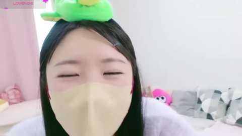 Media: A video of an East Asian woman with long black hair wearing a yellow face mask and a green frog headband, smiling. Background features a pastel room with a pink curtain, a grey and white patterned cushion, and a stuffed toy.