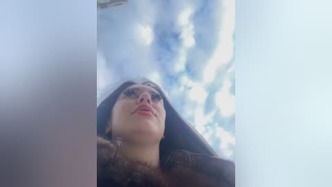 Media: A video of a woman with long dark hair, looking upward through a narrow gap. She has fair skin and a neutral expression. The background shows a bright blue sky with scattered clouds.