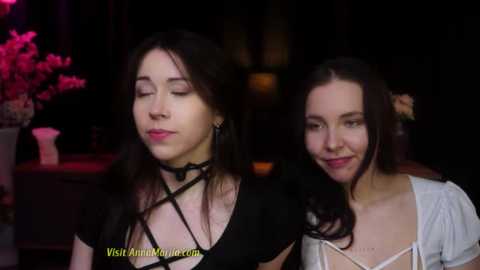 Media: Video of two young women with fair skin, one with dark hair and a black choker, the other with lighter hair and a white top, smiling, dimly lit room with pink flowers in the background.