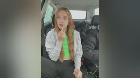 Media: A video of a young woman, likely in her late teens or early twenties, with long blonde hair, sitting in a car. She's wearing a green top and black pants, with a white jacket, and is holding a cigarette.