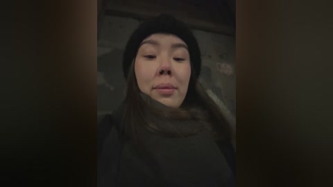 Media: A dimly lit video of a young woman with light skin, long dark hair, and a black beanie, wearing a dark coat, with a serene expression, standing in a gritty urban alley.