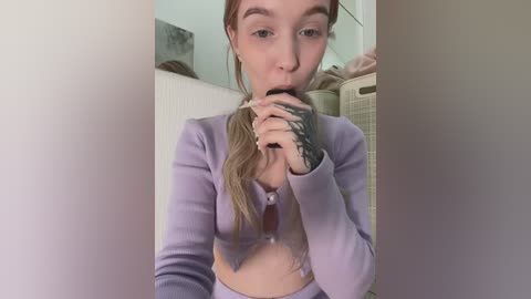 Media: Video of a young woman with fair skin, long brown hair, wearing a lavender cropped cardigan and matching leggings, blowing into a black object, in a laundry room with beige furniture.