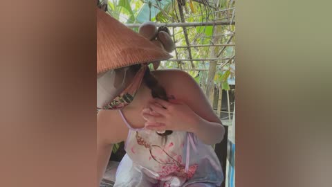 Media: A video of a woman with light skin and brown hair, wearing a floral tank top, kneeling in a bamboo hut. She covers her face with one hand, revealing her bare breasts. The background includes green foliage and a straw hat.