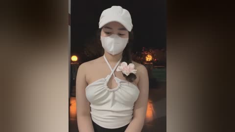 Media: Video of a young woman with medium skin tone and straight black hair, wearing a white halter top, mask, and cap. She stands in a dimly lit, outdoor setting with a blurred background of trees and a lit lantern.