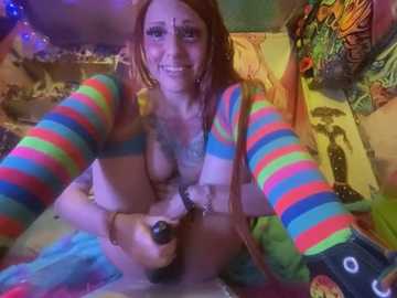 Media: Video of a smiling, topless woman with long red hair, wearing colorful striped sleeves, sitting on a bed in a colorful, messy room with psychedelic posters.