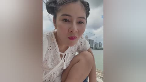 Media: Video of a young Asian woman with fair skin, dark hair styled in buns, wearing a white lace top, sitting near a ferry terminal. Background shows a cloudy sky and Sydney's skyline with the Sydney Harbour Bridge and Opera House.