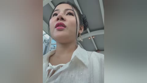 Media: Video of a woman with light brown skin, wearing a white blouse and white gloves, looking up with pink lipstick. She is in an indoor setting with white walls and a ceiling.