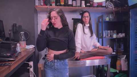 Media: Video of two women in a cluttered kitchen: one in a black crop top and jeans, smoking, the other in a white turtleneck, sitting on a counter.