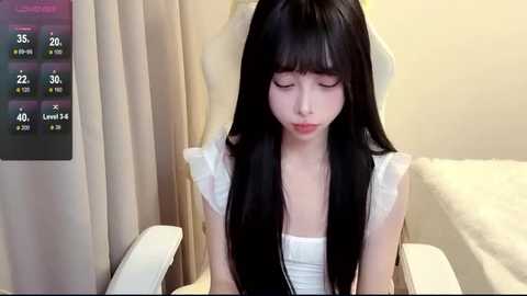 Media: Video of an Asian woman with long black hair and pale skin, wearing a white dress, sitting on a white chair in a beige room with a digital temperature display.