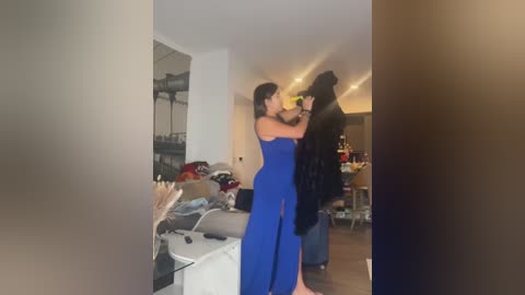 Media: Video of a woman in a blue dress kissing a black-haired man in a modern, cluttered living room with white walls and wooden floors.