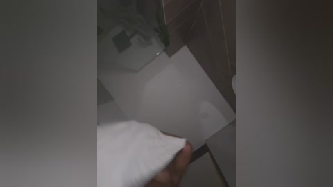 Media: A video showing a close-up view of a hand holding a white paper inside a dimly lit room with beige walls and a partially visible mirror. The image is slightly blurred.