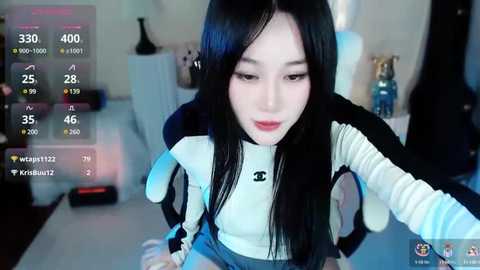 Media: Video of an East Asian woman with long black hair and fair skin, wearing a white and black striped top, sitting on a gaming chair, in a dimly lit room with a TV and keyboard visible.