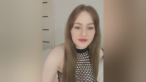 Media: Video of a young Caucasian woman with long, straight brown hair, fair skin, and blue eyes, wearing a black fishnet top, smiling. Background features white wardrobe and beige wall.