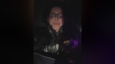 Media: A dimly lit video shows a woman in a black, shiny puffer jacket, with glasses and a partially obscured face. She is sitting in a car, with a purple object visible in the background.