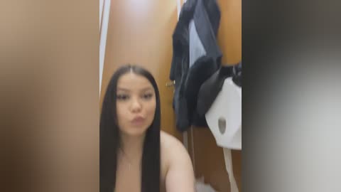 Media: Video of an East Asian woman with long black hair, pouting in a bathroom. Brown walls, a wooden door, and a white paper towel dispenser are visible in the background.