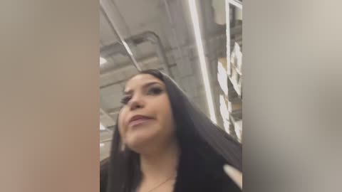 Video of a woman with long black hair and light skin, wearing a black top, captured from a low angle in an industrial setting with exposed pipes and metal shelves in the background.