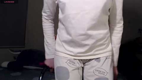 Media: Video of a person wearing a white long-sleeve shirt and white pants with grey patches and text; standing indoors with a dimly lit background.