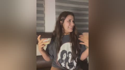 Media: A video of a young woman with long dark hair, wearing a gray Beatles t-shirt, holding a phone, seated on a brown leather couch, with a striped window background.