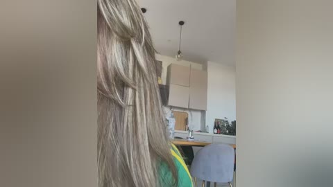 Video of a blonde woman's long, wavy hair partially visible from the side. The background features a modern kitchen with light cabinets, a gray stool, and a wooden countertop.