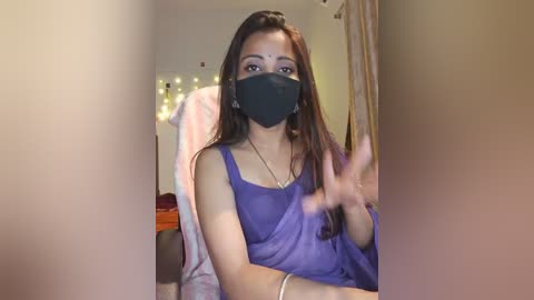 Media: Video of a young South Asian woman with long brown hair, wearing a black face mask, purple dress, and a gold necklace, seated in a cozy room with dim lighting, waving her hand.